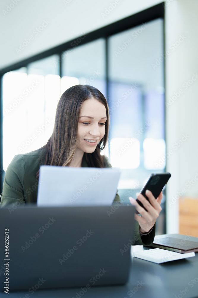 Portrait of a young Asian woman showing a smiling face as she uses his phone, computer and financial documents on her desk in the early morning hours