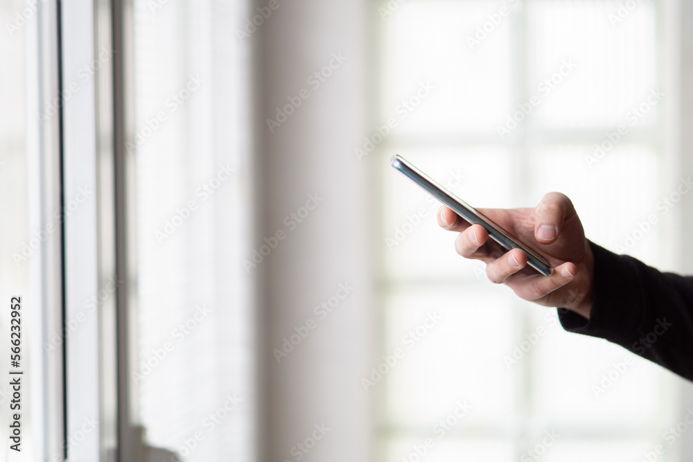 Close up of a man in sweater standing in front of windows at home and holding smartphone. Man hands holding smartphone