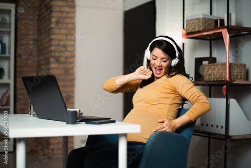 eautiful pregnant listening the music while working on laptop. Young businesswoman working in her office
