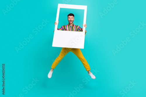 Full body size photo of funky jumping air trampoline man overjoyed wear striped shirt hold paper frame good mood isolated on cyan color background
