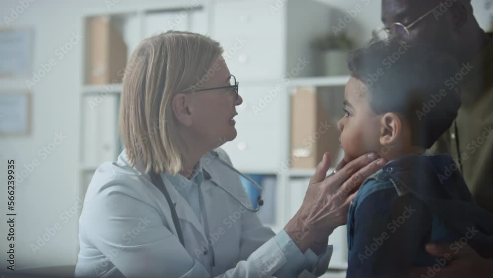 Experienced female pediatrician in eyeglasses and lab coat checking thyroid of little boy and talking to his father during medical consultation in clinics Experienced female pediatrician in eyeglasses and lab coat checking thyroid of little boy and talking to his father during medical consultation in clinics