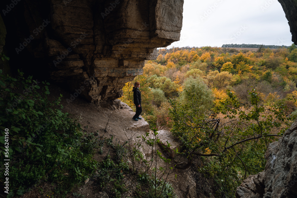 Woman standing by lighted exit in cave, daytime. Atmospheric snapshot in natural rock formations with autumn forest and dramatic gray clouds, tourist attraction. Smotrych river canyon