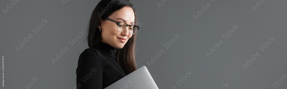 cheerful asian woman in black turtleneck and glasses holding laptop isolated on dark grey, banner cheerful asian woman in black turtleneck and glasses holding laptop isolated on dark grey, banner