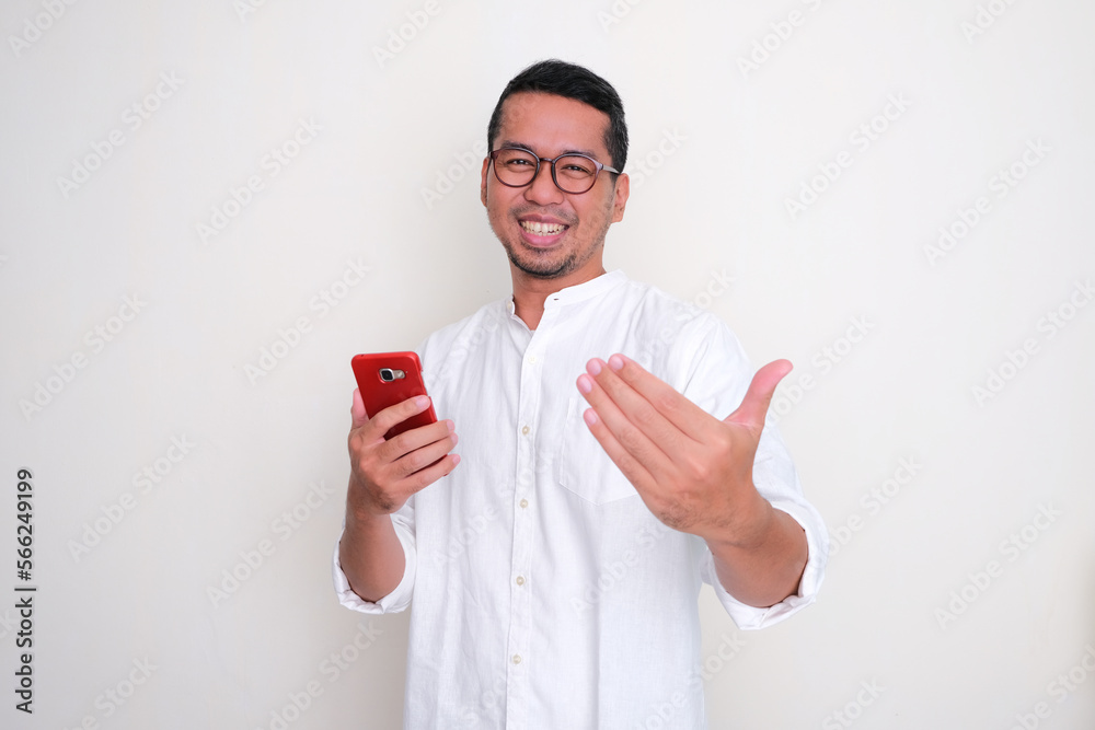 Asian man showing excited expression and doing inviting hand gesture while holding handphone Asian man showing excited expression and doing inviting hand gesture while holding handphone
