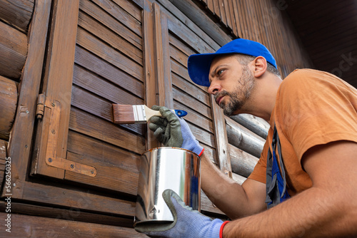 Worker applying protective varnish or wood oil on wooden house cottage exterior walls and window shutters