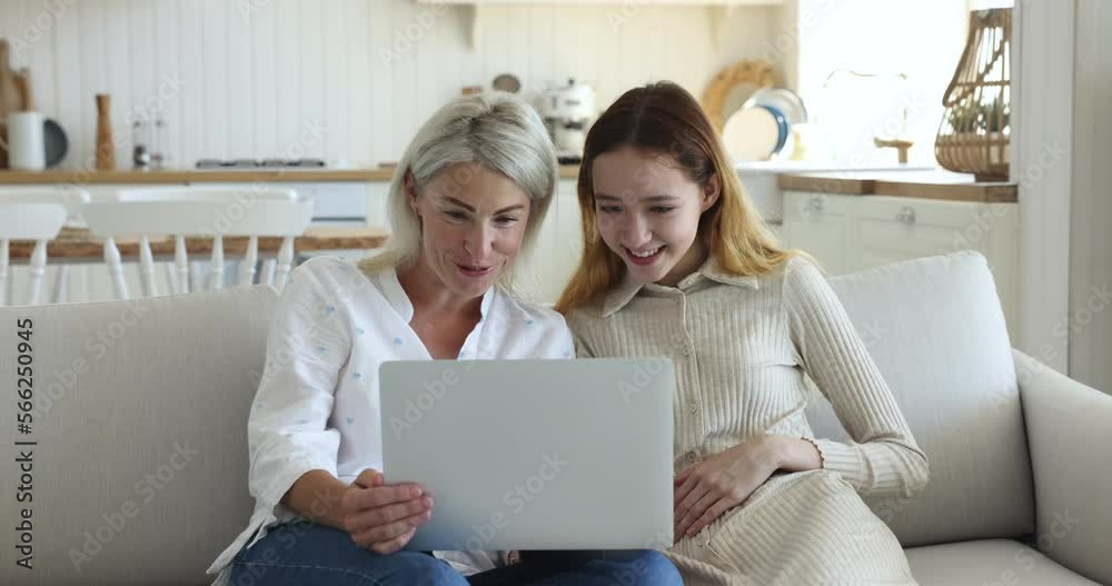 Pretty teenage girl rest on sofa with attractive middle-aged mother with laptop, watch comedy movie or funny vlog content, enjoy video call conversation to friends or family use app. Modern tech, fun