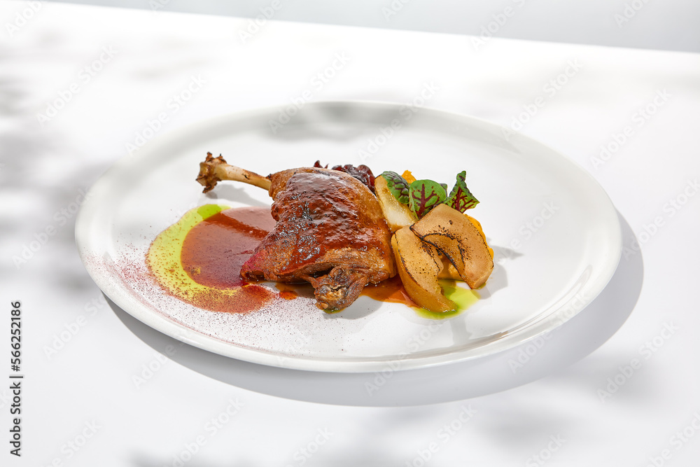 Traditional france dish - duck confit with baked apple on light background with shadows of sunlight. Roasted duck leg with fruits and gravy on white plate in summer dining Elegant roasted leg Traditional france dish - duck confit with baked apple on light background with shadows of sunlight. Roasted duck leg with fruits and gravy on white plate in summer dining Elegant roasted leg