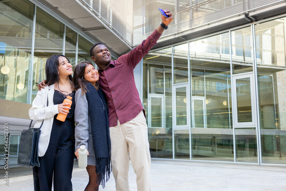 Happy attractive coworkers taking a selfie with a smartphone outside the office. Group of young friends smiling in a work break in the city. Ethnic diversity and social inclusion. Copy space.