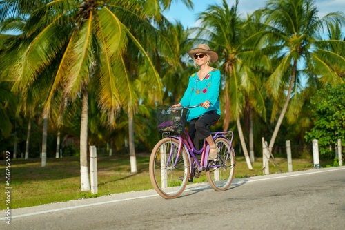 Low angle front view of happy smiling pretty mature senior woman smiling biking towards the camera wearing ethnic clothes and hat and sunglasses on a tropical road.