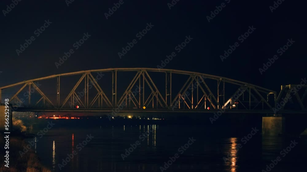 Electric locomotive with freight cars or railway wagon rides on railroad bridge over river at night. Headlights flare reflection in water. Transportation and delivery of cargo in containers between Electric locomotive with freight cars or railway wagon rides on railroad bridge over river at night. Headlights flare reflection in water. Transportation and delivery of cargo in containers between