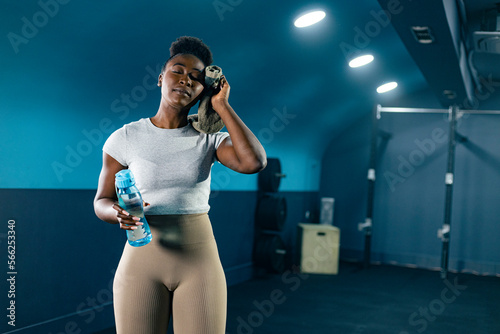Young fit African American woman in sportswear with towel after exercise, holding bottle of water at gym.