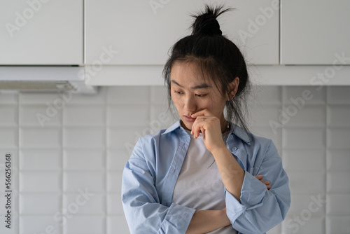 Tableau sur toile Confused apathetic Asian woman touching lips standing in kitchen of own apartment thinking about life goals