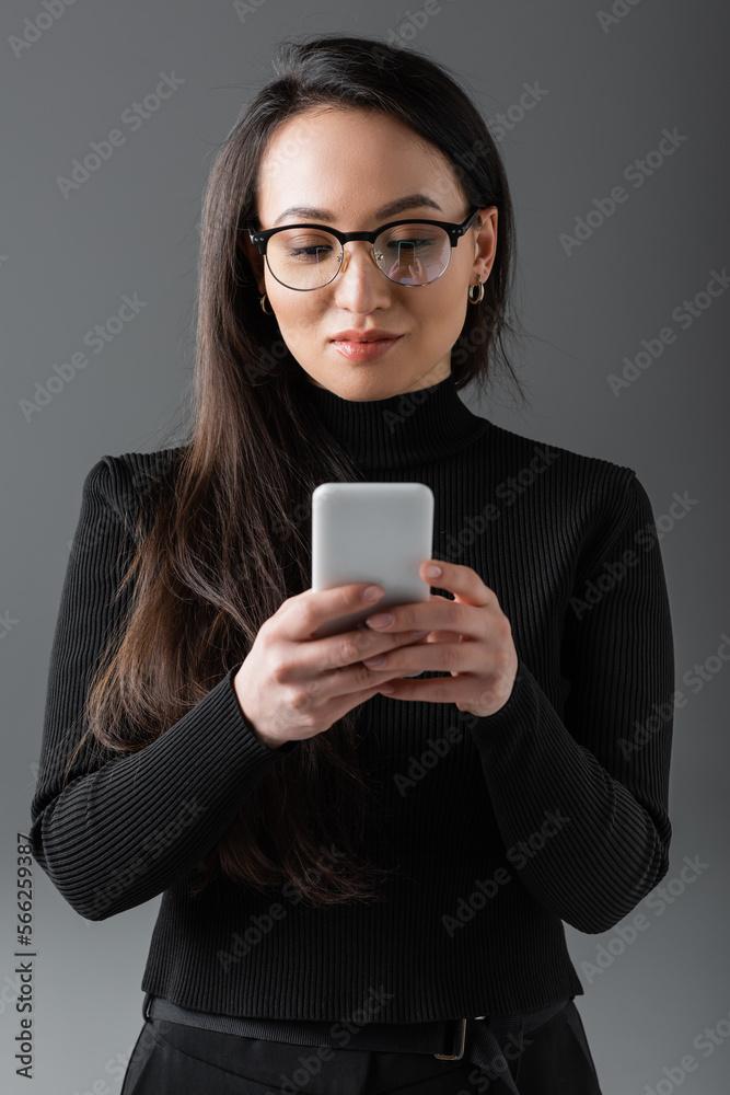 pretty asian woman in black turtleneck and glasses texting on smartphone isolated on dark grey pretty asian woman in black turtleneck and glasses texting on smartphone isolated on dark grey