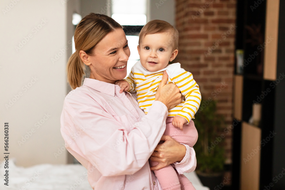 Motherhood concept. Happy mom with infant daughter on arms posing at home, kid looking at camera, free space Motherhood concept. Happy mom with infant daughter on arms posing at home, kid looking at camera, free space