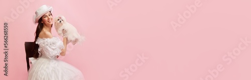 Portrait of beautiful lady, woman in white vintage dress posing with lovely cute dog over pink background. Banner. Concept of 19th century, fashion, comparison of eras, history, retro style photo