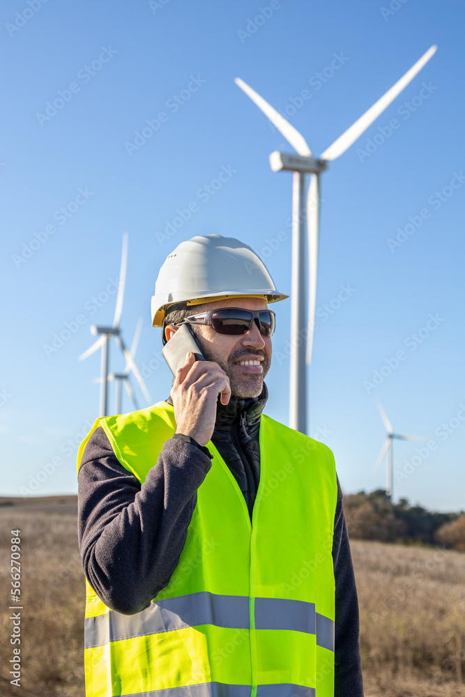 Engineer in protective helmet speaking on smartphone against wind turbines Engineer in protective helmet speaking on smartphone against wind turbines