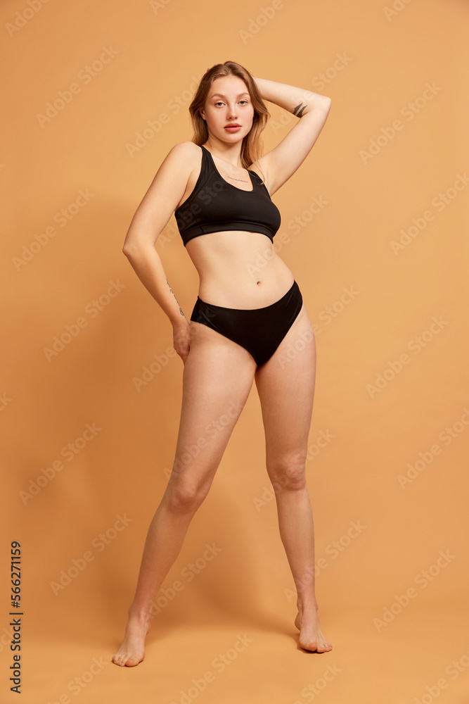 Obraz premium Portrait of young slim beautiful girl wearing black underwear posing over sand color background. Concept of beauty, body and skin care, healthy eating