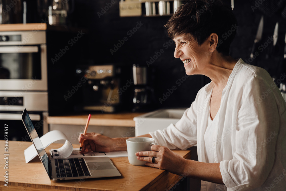 Cheerful senior woman working on laptop computer and writing down notes while sitting in kitchen Cheerful senior woman working on laptop computer and writing down notes while sitting in kitchen