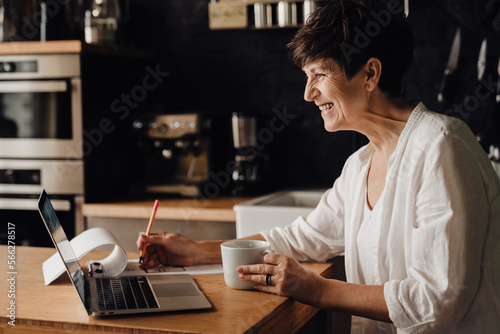 Cheerful senior woman working on laptop computer and writing down notes while sitting in kitchen