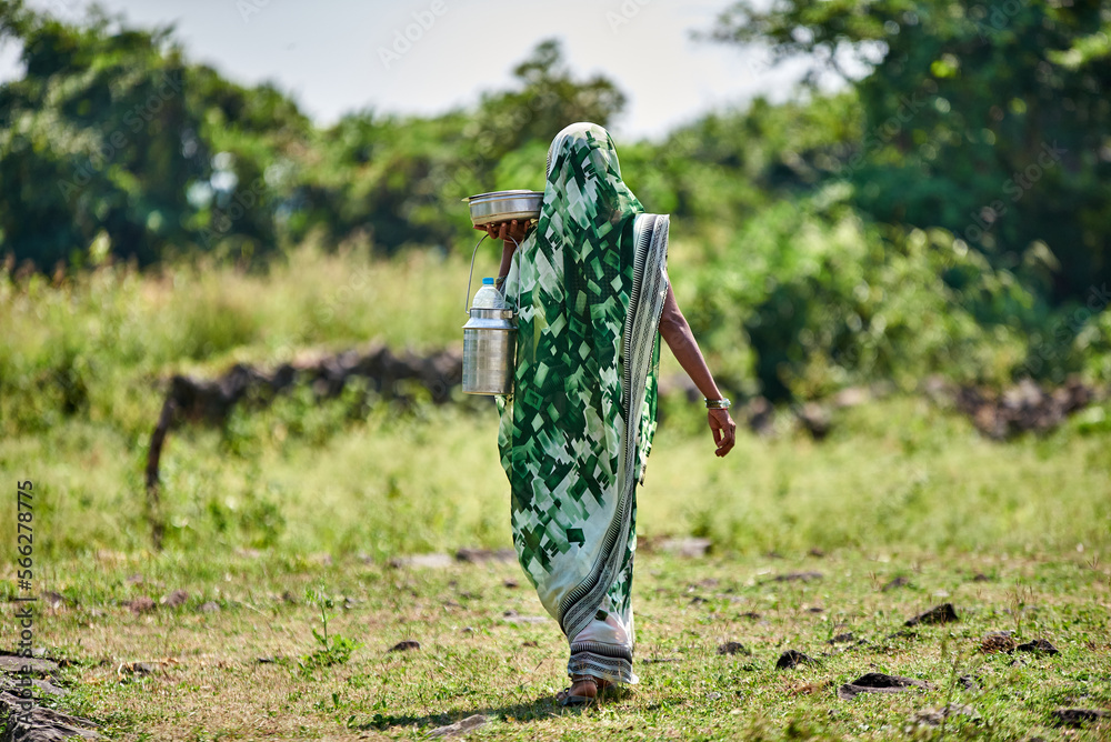 A woman wearing green sari carries pots of food walking through a green village
