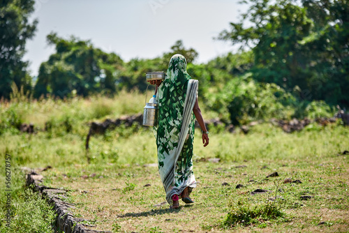 A woman wearing green sari carries pots of food walking through a green village