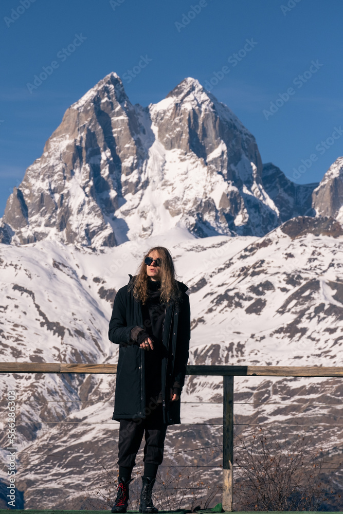 Young stylish man portrait on background of stunning high mountain peaks. Vacation in mountain resort, sunny day, sports and fashion