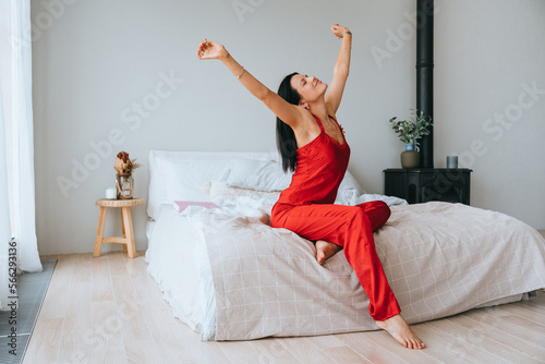 Young brunette hispanic woman in red pyjamas stretching sitting on bed at home on Sunday morning. Satisfied caucasian girl in nightie awakening at hotel room, vacations. Domestic leisure healthy sleep © Iona