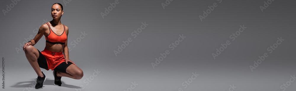 full length of brunette african american sportswoman sitting on haunches on grey background, banner.