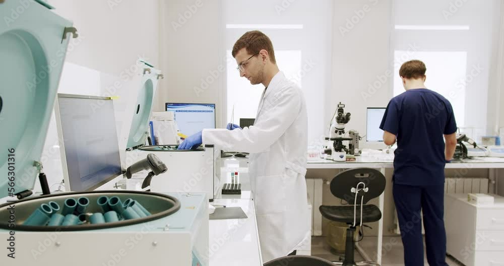 Man working in clinic laboratory. Handheld shot of male medical specialist in uniform preparing samples and using analyzing machine during work in lab of modern hospital. Man working in clinic laboratory. Handheld shot of male medical specialist in uniform preparing samples and using analyzing machine during work in lab of modern hospital.