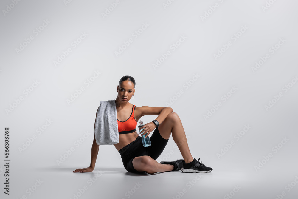 Obraz premium young african american woman in sports bra and bike shorts sitting with sports bottle and towel on grey background.