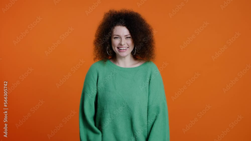 Cheerful stunning young woman with stylish afro hairstyle, waves hello with hand on color background