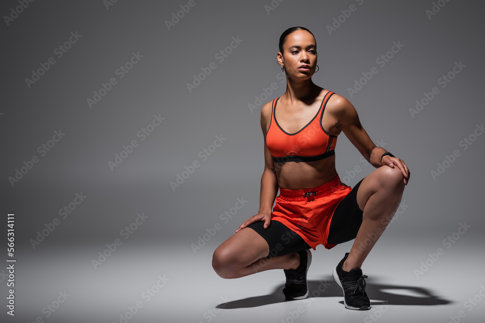 full length of brunette african american sportswoman sitting on haunches on grey background. full length of brunette african american sportswoman sitting on haunches on grey background.