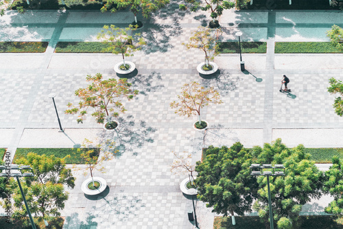 A drone point-of-view shot with a park street treelined with different plants and trees; tiles and stripes of lawn on the ground, a single motion-blurred silhouette of a man riding an e-scooter