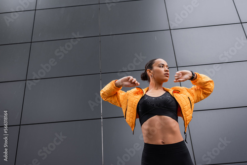 Fotografie low angle view of brunette african american sportswoman in sports bra and yellow puffer jacket exercising outside