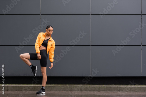 full length of brunette african american sportswoman in bike shorts and yellow puffer jacket posing outside.