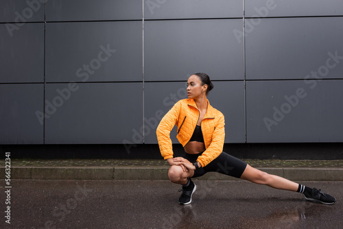 full length of stylish african american sportswoman in bike shorts and yellow puffer jacket stretching near grey building.