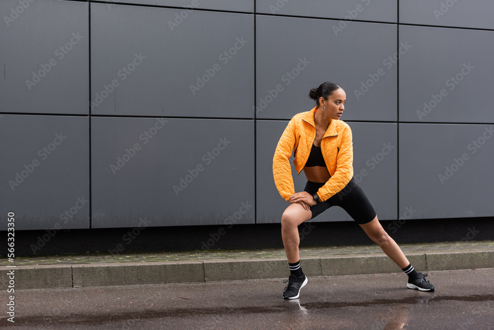 full length of brunette african american sportswoman in bike shorts and yellow puffer jacket exercising outside.