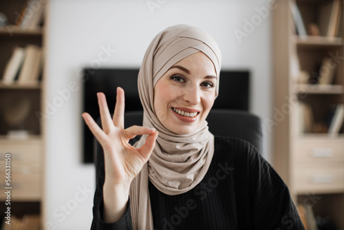 Smiling positive confident muslim woman in formal wear and hijab sitting at office chair showing sign ok. Company worker with headscarf looking at camera indoors. Concept of people and business.