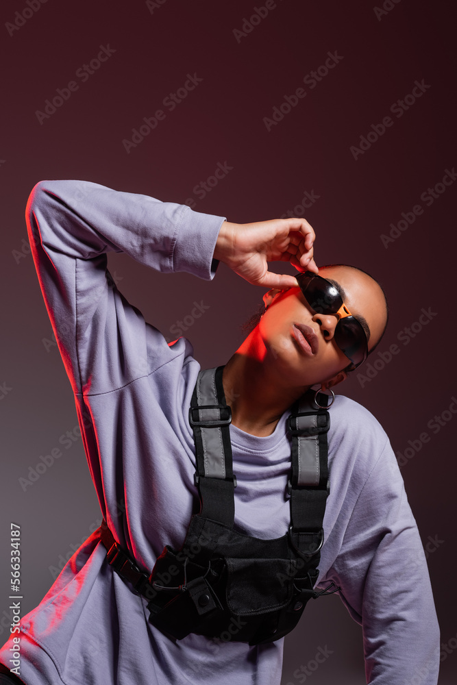 portrait of african american woman in sweatshirt adjusting trendy sunglasses on grey with red light.