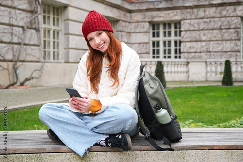 Redhead girl, female student sits with mobile phone on bench in parj, leans on her backpack. Woman browsing social media app feed on her smartphone photo