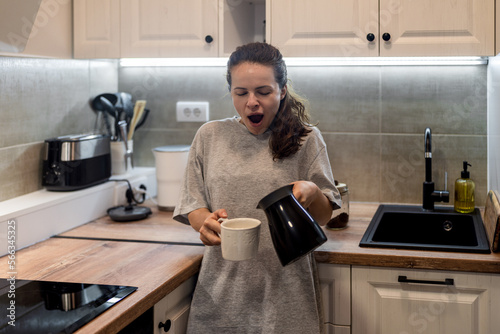 Morning lifestyle woman standing in the kitchen pouring coffee into mug from turkish coffee maker and yawning.