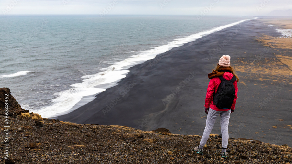 Beautiful girl in pink jacket and backpack looks at vast empty black sand beach Dyrhólaey and atlantic ocean iceland, europe