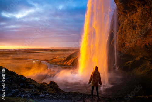 Girl in yellow raincoat admires the stunning colorful sunset spectacle from behind the impressive Seljalandsfoss waterfall. Iceland - Golden Circle