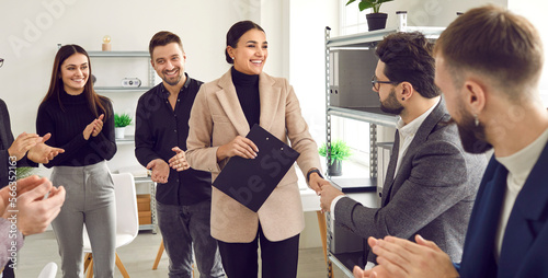 Team of people standing in office, clapping, and congratulating young woman on promotion. Male boss shakes hands with female employee thanking her for help and good work. Business, recognition concept