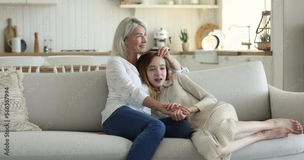 Loving 45s mum hugs, strokes, touch hair while talk to teenage 13s daughter, resting together on cozy couch at home spend carefree leisure, lead pleasant conversation, enjoy harmonic friendly relation