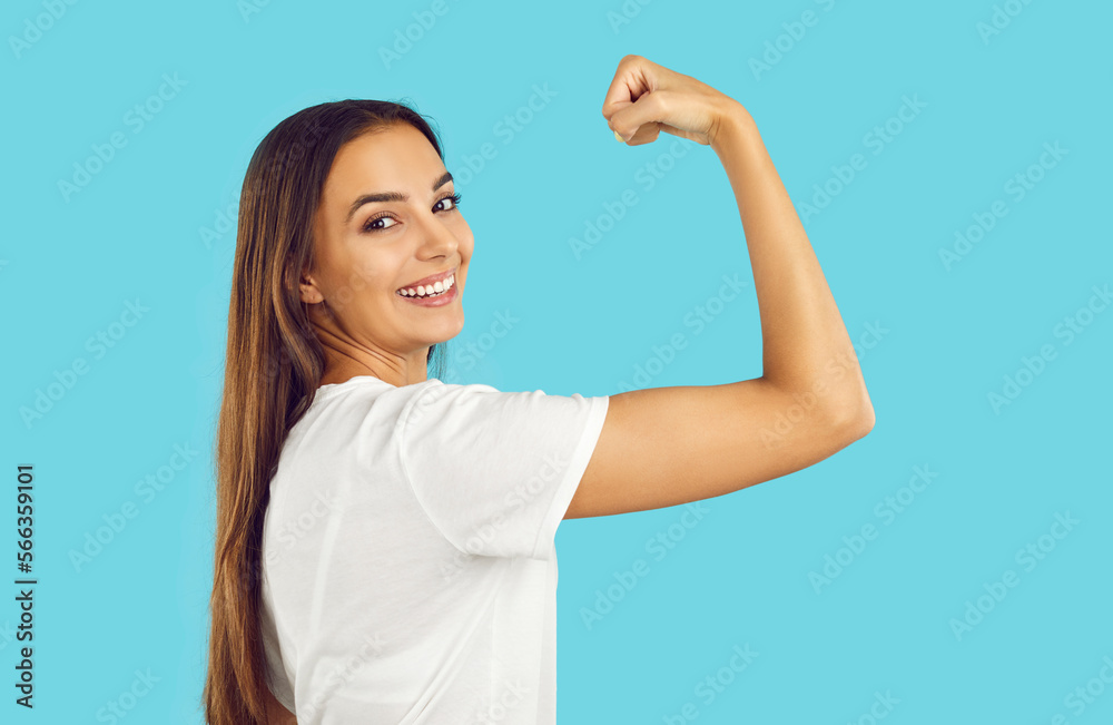 Confident strong happy woman with long hair smiling and showing her power flexing arm on blue background looking at camera. Girl power, strength, independence. Strong gesture with clenching fist. Confident strong happy woman with long hair smiling and showing her power flexing arm on blue background looking at camera. Girl power, strength, independence. Strong gesture with clenching fist.
