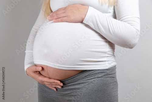 A pregnant girl in a white jacket, hugs her stomach with her hands.