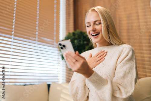 Close up of laughing young woman receiving exciting message on smartphone, reading text in screen, smiling, laughing, sitting on couch, using online virtual app on mobile phone at home.