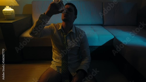 Upset Alcoholic Man Without Trousers in a White Shirt and Tie Sits on the Floor of the House and Gets Drunk with Alcohol from a Glass Bottle at Night. Concept of Alcoholism and Depression.