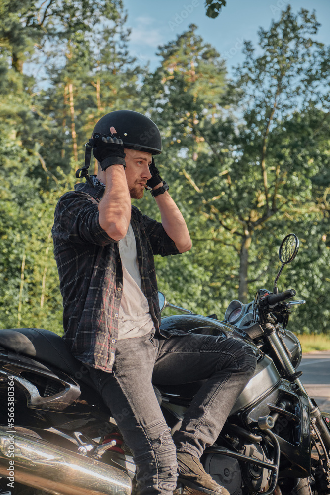 A young male biker travels on a motorcycle alone, stopped and puts on a helmet on the side of a forest road